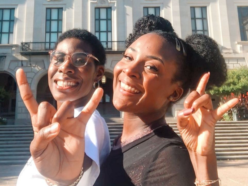 two African-American female students doing hook'em horns in front of the Gregory Gymnasium on UT Campus